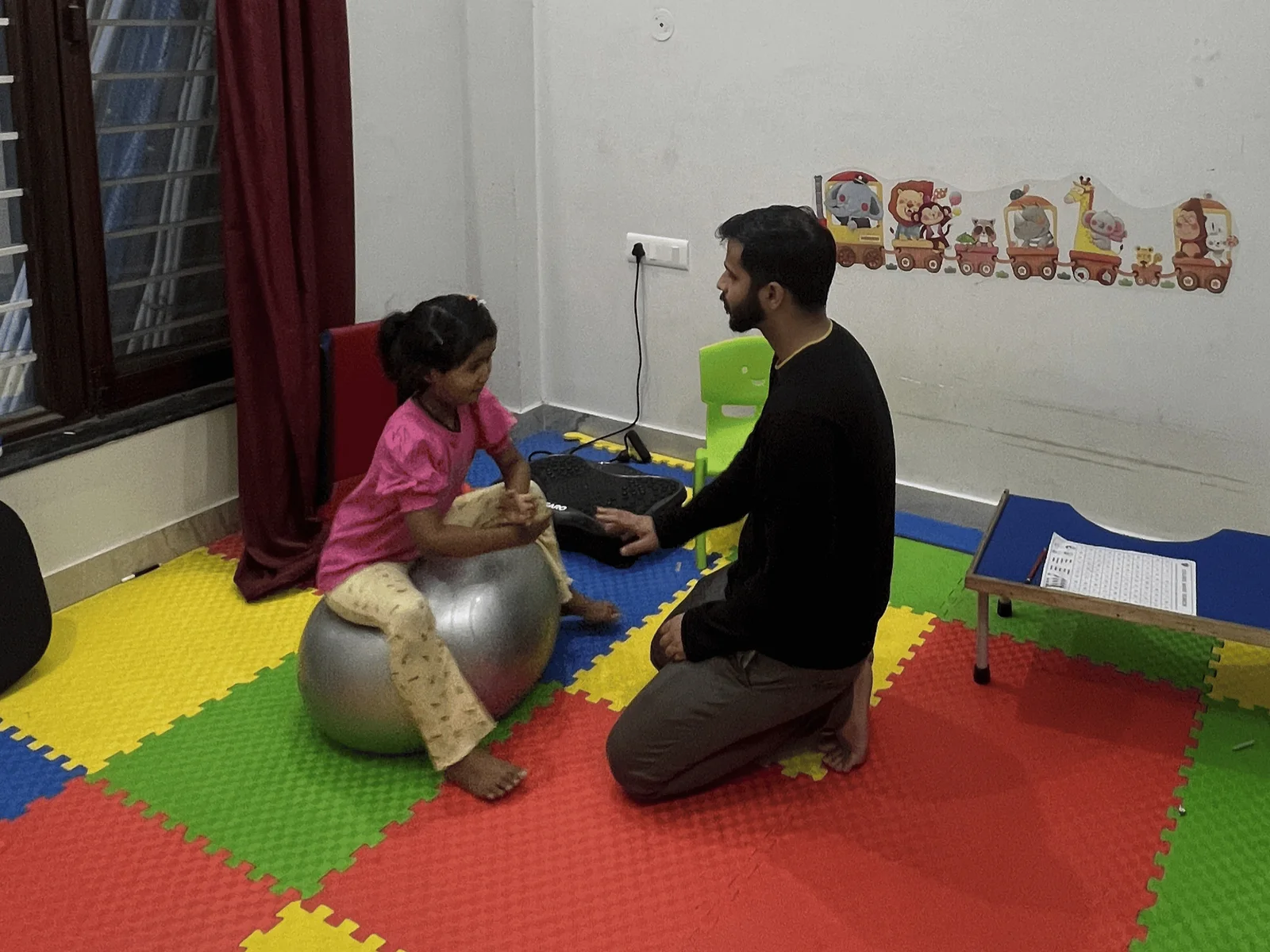 A therapist kneeling on a play mat while a child balances on a therapy ball