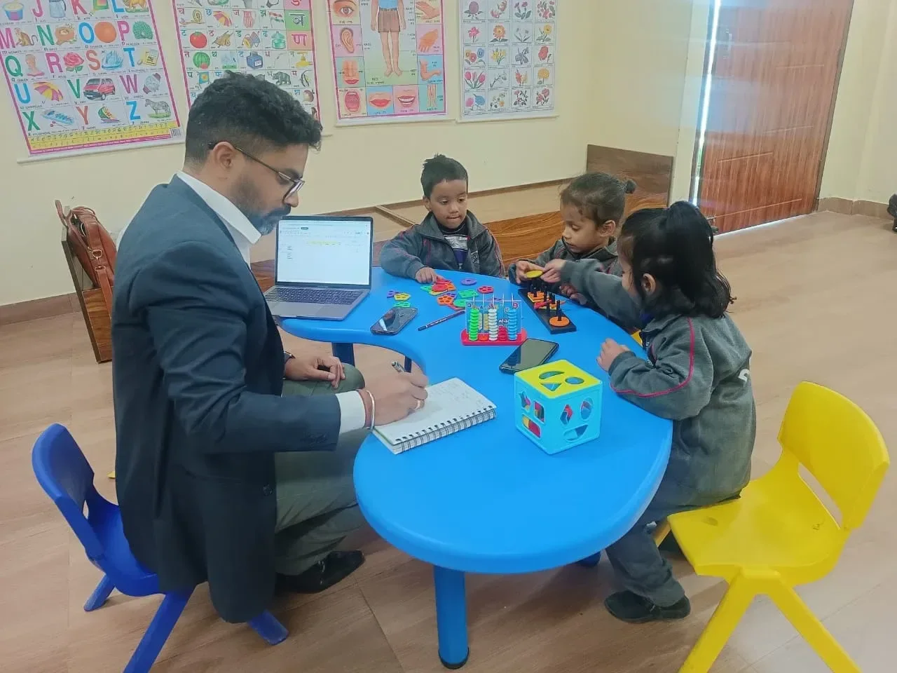 Rishi Kant Upadhyay seated at a low table with three children working on colour and shape activities, a laptop and notebook in front of him